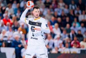 Ein männlicher Handballspieler in einem weiß-goldenen Trikot bereitet sich während eines Spiels auf den Wurf vor, während im Hintergrund eine unscharfe Menschenmenge zusieht.