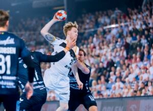 Ein Handballspieler in einem weißen Trikot springt, um den Ball zu werfen, während Spieler in dunklen Trikots versuchen, ihn zu blockieren. Das Spiel findet in einer Halle statt, die mit einer jubelnden Menge gefüllt ist.