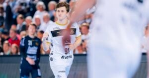 Ein männlicher Handballspieler in einem weißen Trikot joggt auf dem Spielfeld, mit einem verschwommenen Mannschaftskameraden im Vordergrund und Zuschauern im Hintergrund. Die Szene ist hell und actionbetont.