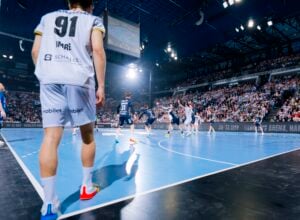 Ein Handballspiel in einer großen Halle mit vielen Zuschauern. Spieler in weißen und marineblauen Trikots sind in Aktion, während ein Spieler in einem weißen Trikot mit der Nummer 91 im Vordergrund steht und das Spiel beobachtet.