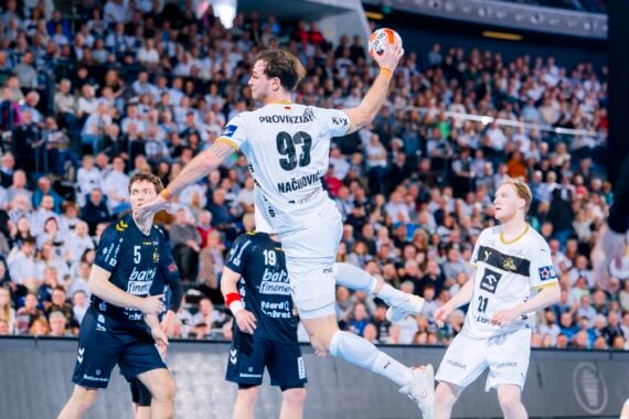 Ein Handballspieler in einem weißen Trikot springt in die Luft, um den Ball zu werfen, umgeben von Spielern in dunklen Trikots, mit einem großen, jubelnden Publikum im Hintergrund.