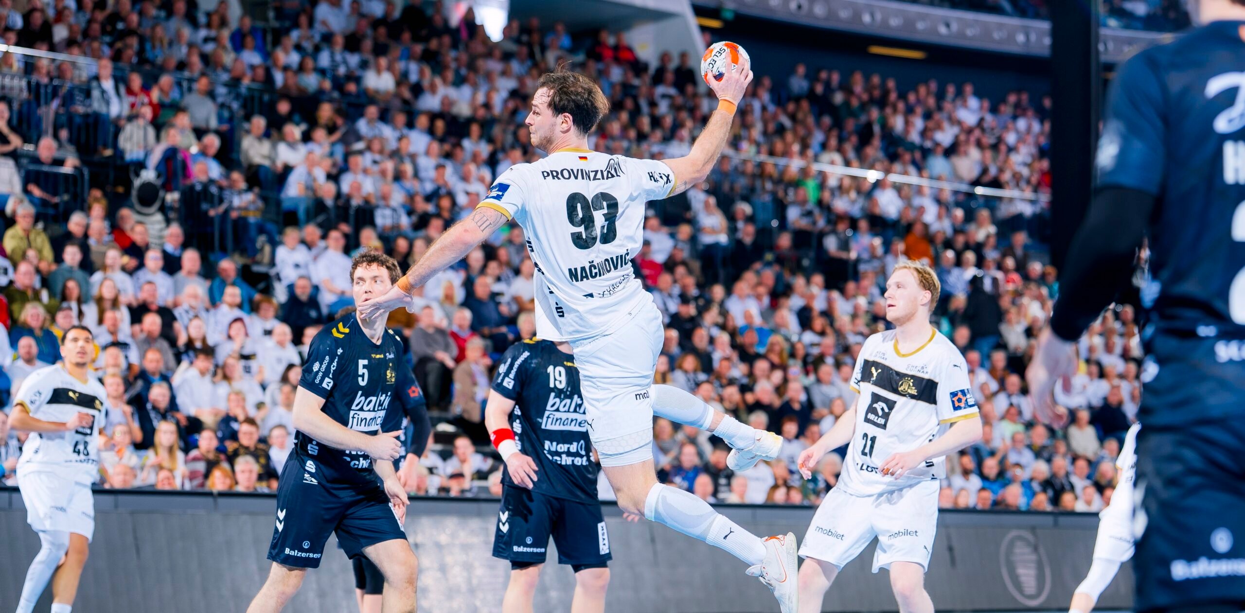 Ein Handballspieler in einem weißen Trikot springt in die Luft, um den Ball zu werfen, umgeben von Spielern in dunklen Trikots, mit einem großen, jubelnden Publikum im Hintergrund.