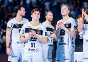 Eine Gruppe männlicher Handballspieler in weißen und schwarzen Trikots steht lächelnd und klatschend auf einem Spielfeld, während im Hintergrund eine unscharfe Menschenmenge zu sehen ist.
