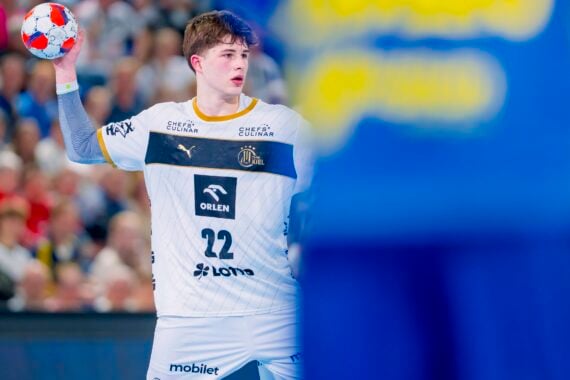 Ein männlicher Handballspieler in einem weißen Trikot mit der Nummer 22 hält einen roten und blauen Ball und bereitet sich auf den Wurf vor, während im Vordergrund ein unscharfer Gegner in Blau erscheint. Im Hintergrund ist eine überfüllte Arena zu sehen.