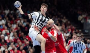 Ein Handballspieler in einem schwarz-weißen Trikot springt, um den Ball zu werfen, während ein Verteidiger in einem roten Trikot versucht, ihn zu blockieren. Das Spiel findet in einer überfüllten Halle mit Zuschauern im Hintergrund statt.