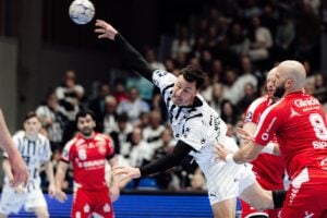 Ein Handballspieler in einem weiß-schwarzen Trikot springt, um den Ball zu werfen, während Spieler in roten Trikots versuchen, ihn während eines intensiven Spiels zu blockieren, mit einem verschwommenen Publikum im Hintergrund.