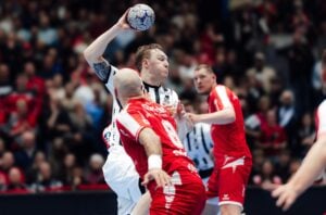 Ein Handballspieler in einem weißen Trikot bereitet sich darauf vor, den Ball zu werfen, während er von Spielern in roten Trikots während eines Spiels eng verteidigt wird.