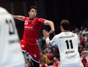 Ein männlicher Handballspieler in einem roten Trikot springt, um den Ball zu werfen, während er von einem Spieler in einem weißen Trikot verteidigt wird. Die Aktion findet in einer Halle statt, mit einem unscharfen Publikum im Hintergrund.