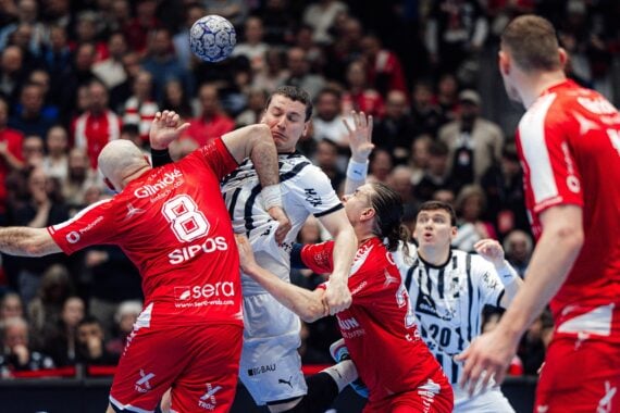 Mehrere Handballspieler in rot-weißen Trikots springen und greifen während eines spannungsgeladenen Spiels nach dem Ball, während im Hintergrund ein dicht gedrängtes Publikum zusieht.