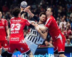 Ein Handballspieler in einem weiß-schwarzen Trikot versucht zu schießen, während er von zwei Verteidigern in roten Trikots bei einem spannenden Spiel in einer überfüllten Halle geblockt wird.