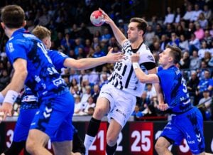 Ein Handballspieler in einem weiß-schwarzen Trikot springt, um den Ball zu werfen, während drei gegnerische Spieler in blauen Trikots versuchen, ihn während eines Spiels in einer überfüllten Halle zu blockieren.