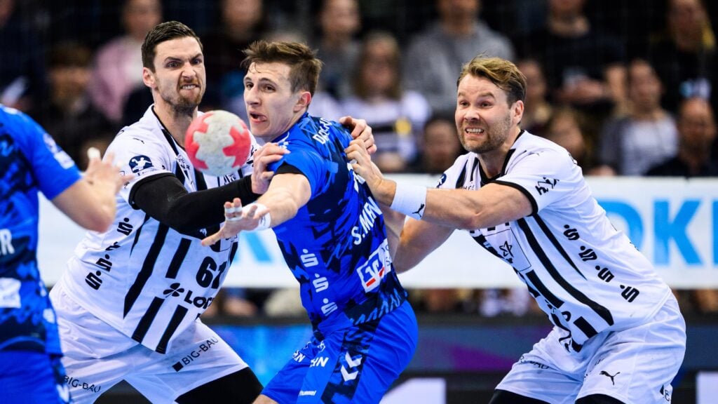 Drei männliche Handballspieler liefern sich einen intensiven Wettkampf; ein Spieler in einem blauen Trikot versucht, den Ball zu passen, während zwei Spieler in weißen Trikots ihn eng verteidigen, inmitten einer verschwommenen Menschenmenge im Hintergrund.