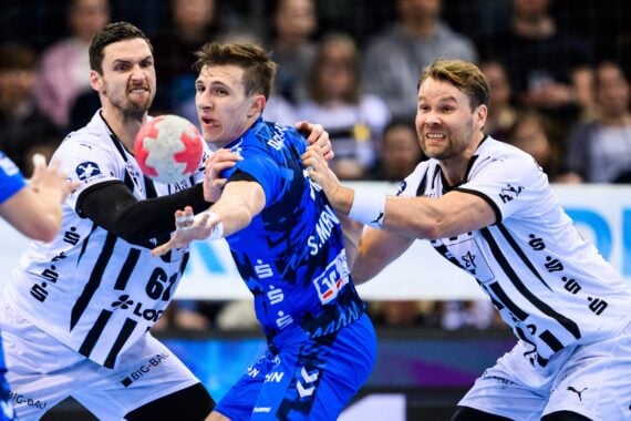 Drei männliche Handballspieler liefern sich einen intensiven Wettkampf; ein Spieler in einem blauen Trikot versucht, den Ball zu passen, während zwei Spieler in weißen Trikots ihn eng verteidigen, inmitten einer verschwommenen Menschenmenge im Hintergrund.