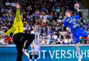 Ein Handballspieler in einem blauen Trikot springt, um den Ball in Richtung Tor zu werfen, während der Torwart in Gelb versucht, ihn zu blocken. Auf den Tribünen im Hintergrund sitzen Zuschauer, die das intensive Spiel verfolgen.