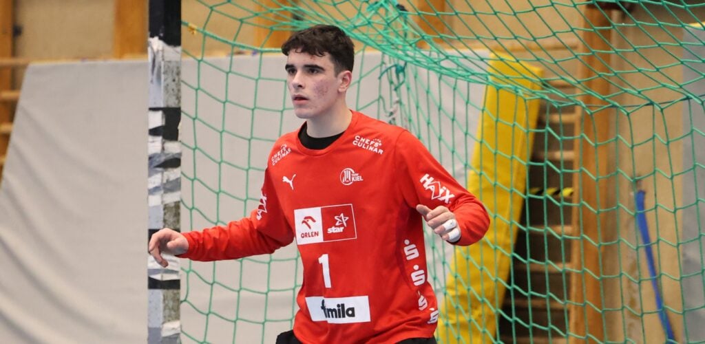 26_pm_Dengler_Titel3 Ein männlicher Handballtorwart in einem roten Trikot mit der Nummer 1, der den THW Kiel Handball-Bundesliga Saison 2025/2026 repräsentiert, steht wachsam vor dem Tor, bereit, einen Schuss zu blocken. Im Hintergrund sind ein grünes Netz und eine Sporthalle zu sehen.