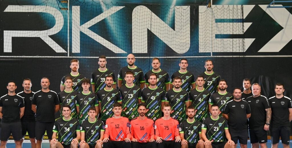 Eine Herren-Handballmannschaft posiert in einer Halle für ein Gruppenfoto vor einem großen RK NEXE-Wandschild. Die Spieler tragen schwarze und grüne Trikots, vorne sitzen zwei Torhüter in orangefarbenen Trikots. Die Trainer stehen auf beiden Seiten.