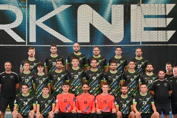 Eine Herren-Handballmannschaft posiert in einer Halle für ein Gruppenfoto vor einem großen RK NEXE-Wandschild. Die Spieler tragen schwarze und grüne Trikots, vorne sitzen zwei Torhüter in orangefarbenen Trikots. Die Trainer stehen auf beiden Seiten.