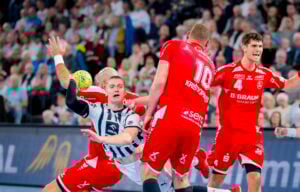 Ein Handballspieler in einem weiß-blauen Trikot bereitet sich im Fallen auf einen Schuss vor, umgeben von drei Verteidigern in roten Trikots während eines intensiven Spiels, während die Zuschauer im Hintergrund zusehen.