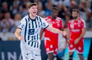Ein männlicher Handballspieler in einem weiß-schwarz gestreiften Trikot jubelt mit einer geballten Faust auf dem Spielfeld, während im Hintergrund gegnerische Spieler in roten Trikots zu sehen sind.