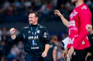 Ein lächelnder Trainer im schwarzen Sportdress mit Schlüsselband und HAHN auf dem Trikot pumpt auf dem Handballfeld während des Vorberichts der THW Kiel Handball-Bundesliga Saison 2025/2026 gegen den TVB Stuttgart jubelnd die Faust, dahinter verschwommene Spieler und Zuschauer.