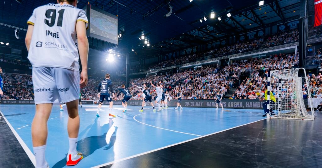 HANDBALL 2025/26: THW Kiel - SG Flensburg-Handewitt Ein Handballspiel in einer überfüllten Sporthalle. Spieler in weißen und marineblauen Trikots sind auf dem Spielfeld, ein weiß gekleideter Spieler steht im Vordergrund und die Zuschauer füllen die Tribünen im Hintergrund.