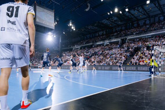 Ein Handballspiel in einer überfüllten Sporthalle. Spieler in weißen und marineblauen Trikots sind auf dem Spielfeld, ein weiß gekleideter Spieler steht im Vordergrund und die Zuschauer füllen die Tribünen im Hintergrund.
