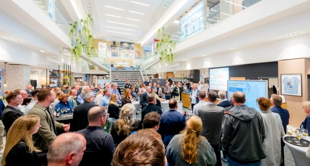 Eine große Gruppe von Menschen versammelt sich in einem Innenraum und hört sich eine Präsentation an. Der Raum ist modern und gut beleuchtet, mit Glasgeländern, von oben herabhängenden Grünpflanzen und einer Treppe im Hintergrund.