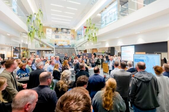 Eine große Gruppe von Menschen versammelt sich in einem Innenraum und hört sich eine Präsentation an. Der Raum ist modern und gut beleuchtet, mit Glasgeländern, von oben herabhängenden Grünpflanzen und einer Treppe im Hintergrund.