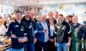 Eine Gruppe von Menschen, einige in schwarzen Sportjacken und einer in einer grünen Schürze, posiert und lächelt zusammen in einer hellen, geschäftigen Innenumgebung, in der im Hintergrund Lebensmittel und Küchengeräte zu sehen sind.