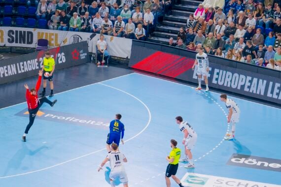 Ein Handballspieler in Blau bereitet sich darauf vor, einen Elfmeter zu schießen, während der Torwart in Rot bereitsteht. Die Spieler in Weiß und die Schiedsrichter schauen zu, während im Hintergrund ein großes Publikum und Banner der European League 2023 zu sehen sind.