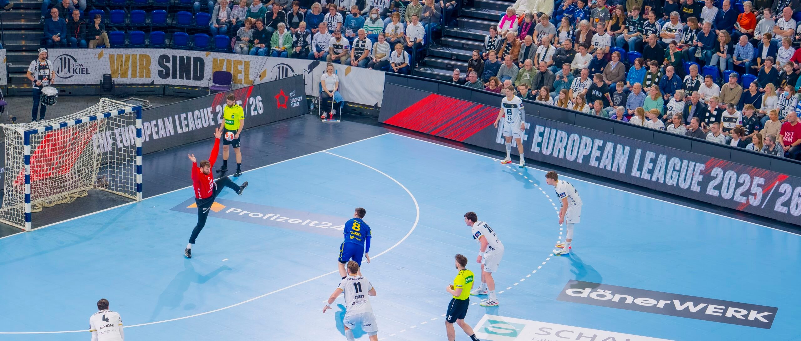 Ein Handballspieler in Blau bereitet sich darauf vor, einen Elfmeter zu schießen, während der Torwart in Rot bereitsteht. Die Spieler in Weiß und die Schiedsrichter schauen zu, während im Hintergrund ein großes Publikum und Banner der European League 2023 zu sehen sind.