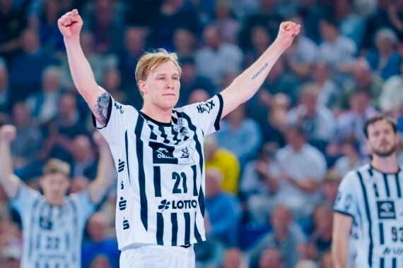 Ein Handballspieler des THW Kiel im schwarz-weiß gestreiften Trikot reißt während der Handball-Bundesliga Saison 2025/2026 beide Arme jubelnd in die Höhe. Im Hintergrund sind zwei Mitspieler und eine unscharfe Menschenmenge zu sehen.