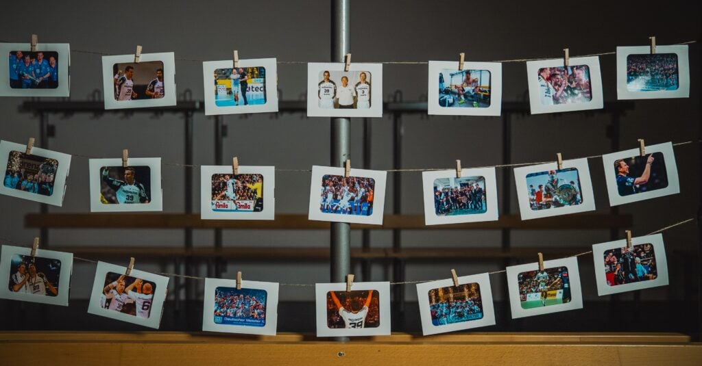 An zwei Schnüren sind mit Wäscheklammern Fotos im Polaroid-Stil befestigt, die verschiedene Gruppen- und Sportszenen zeigen und in Innenräumen vor einem verschwommenen Hintergrund mit Holzbänken hängen.