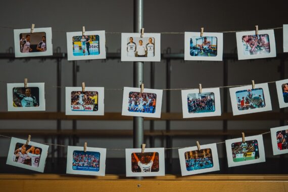 An zwei Schnüren sind mit Wäscheklammern Fotos im Polaroid-Stil befestigt, die verschiedene Gruppen- und Sportszenen zeigen und in Innenräumen vor einem verschwommenen Hintergrund mit Holzbänken hängen.