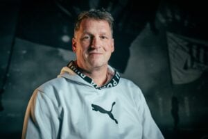 Ein Mann mittleren Alters mit kurzen Haaren und einem weißen Puma-Kapuzenpullover sitzt da und lächelt sanft in die Kamera. Der Hintergrund ist dunkel und unscharf und zeigt undeutliche Formen oder Flaggen.