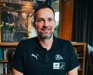 Ein Mann mit kurzen dunklen Haaren und einem Bart, der ein schwarzes Poloshirt mit Team Kiel-, Puma-, Orlen- und Star-Logos trägt, sitzt in einem Raum mit Bücherregalen und Blumen im Hintergrund und lächelt in die Kamera.