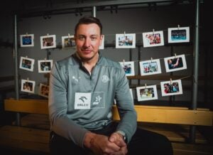 Ein Mann in einer grauen Sportjacke sitzt auf einer Holzbank und lächelt leicht. Hinter ihm hängen Fotos an Schnüren in einem Regal, die verschiedene bunte Momente und Ereignisse zeigen. Der Hintergrund ist schummrig beleuchtet.