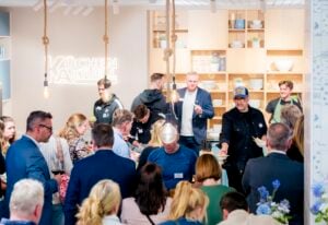 Eine Gruppe von Personen nimmt an einer lebhaften Kochvorführung in einem modernen Küchenausstellungsraum teil, bei der Köche Speisen zubereiten und Gäste zusehen und Gerichte probieren. Im Hintergrund sind Regale mit Geschirr und Küchenutensilien zu sehen.