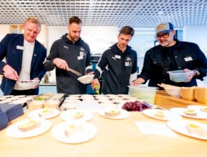 In einem hell erleuchteten Innenraum stehen vier Männer mit Namensschildern und legerer Kleidung an einem Tisch und stellen Essen in Schüsseln und Tellern zusammen oder servieren es.