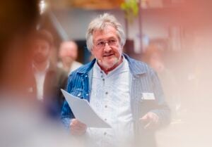 Ein älterer Mann mit grauem Haar und Brille, der eine gestreifte blaue Jacke und ein weißes Hemd trägt, spricht und hält Papiere in der Hand. Er hat ein Mikrofon-Headset und steht in einem Innenraum, mit unscharfen Menschen im Hintergrund.