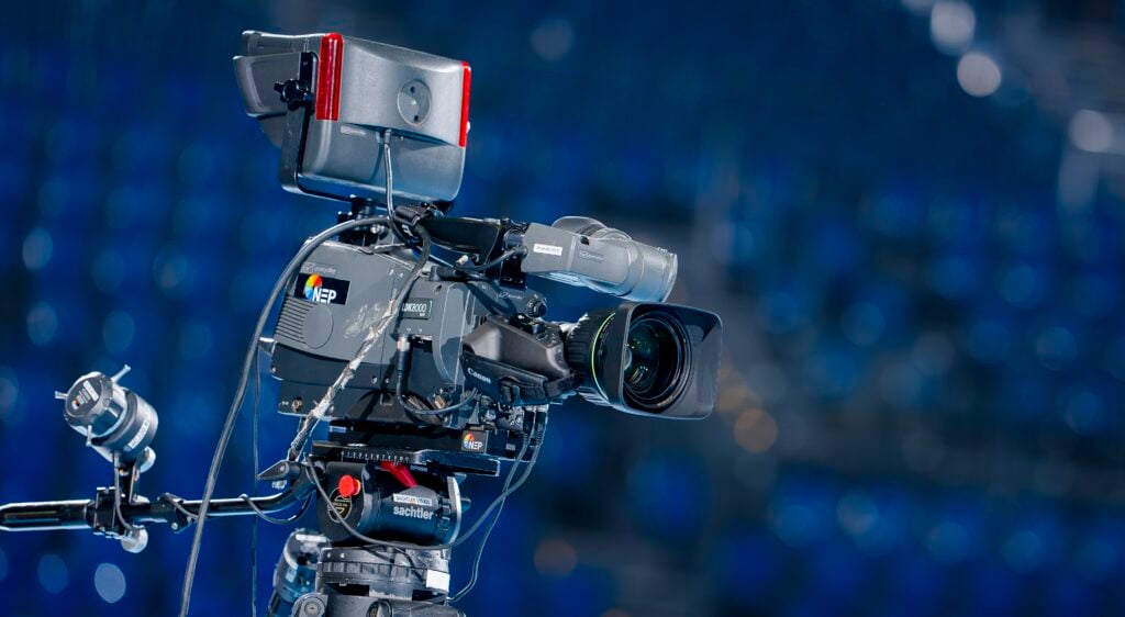 Eine professionelle Fernsehkamera mit verschiedenen Aufsätzen ist auf einem Stativ aufgebaut, fokussiert und bereit für die Aufnahme. Der Hintergrund ist in Blautönen gehalten, was auf eine Innenveranstaltung oder ein Studio hindeutet.