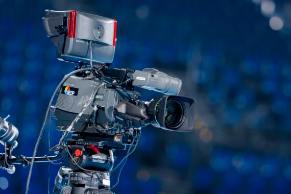 Eine professionelle Fernsehkamera mit verschiedenen Aufsätzen ist auf einem Stativ aufgebaut, fokussiert und bereit für die Aufnahme. Der Hintergrund ist in Blautönen gehalten, was auf eine Innenveranstaltung oder ein Studio hindeutet.