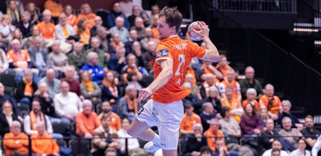 Ein männlicher Handballspieler in einem orangefarbenen Trikot springt in die Luft, um den Ball während eines Spiels zu werfen, während im Hintergrund eine Menge von Zuschauern von der Tribüne aus zusieht.