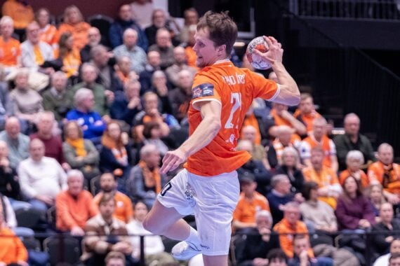 Ein männlicher Handballspieler in einem orangefarbenen Trikot springt in die Luft, um den Ball während eines Spiels zu werfen, während im Hintergrund eine Menge von Zuschauern von der Tribüne aus zusieht.