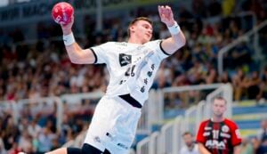 Ein Handballspieler in einem weißen Trikot springt während eines Spiels zum Wurf, während im Hintergrund eine Menschenmenge zusieht und ein gegnerischer Spieler in einem schwarz-roten Trikot in der Nähe steht.