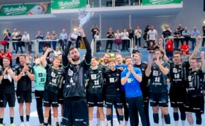 Eine Männer-Handballmannschaft in schwarzen Trikots feiert, während ein Spieler einen Pokal hochhält. Die Mannschaftskameraden jubeln und klatschen, während im Hintergrund Zuschauer zusehen und Fotos machen.