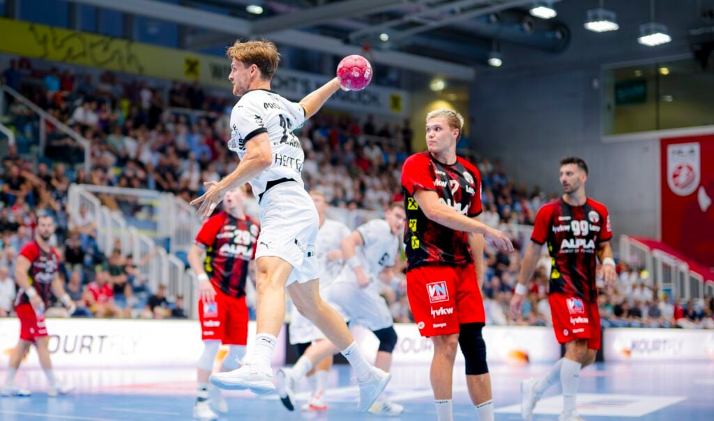 Ein Handballspieler in einem weißen Trikot springt, um den Ball zu werfen, während Spieler in roten und schwarzen Trikots verteidigen. Das Spiel findet in einer überfüllten Halle mit Zuschauern im Hintergrund statt.