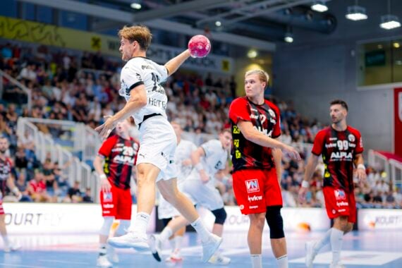 Ein Handballspieler in einem weißen Trikot springt, um den Ball zu werfen, während Spieler in roten und schwarzen Trikots verteidigen. Das Spiel findet in einer überfüllten Halle mit Zuschauern im Hintergrund statt.