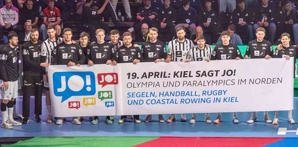 Eine Herren-Handballmannschaft in schwarz-weißen Trikots steht auf einem Spielfeld und hält ein Banner, das für die Olympischen Spiele und Paralympics in Norddeutschland wirbt, mit Text über Segeln, Handball, Rugby und Rudern in Kiel.