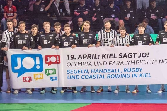 Eine Herren-Handballmannschaft in schwarz-weißen Trikots steht auf einem Spielfeld und hält ein Banner, das für die Olympischen Spiele und Paralympics in Norddeutschland wirbt, mit Text über Segeln, Handball, Rugby und Rudern in Kiel.